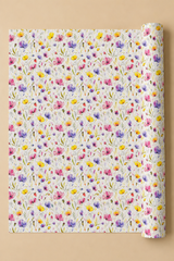 A roll of wrapping paper with a dense, colorful watercolor floral pattern on a white background, partially unrolled and displayed against a solid beige background.