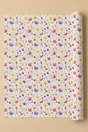 A roll of wrapping paper with a dense, colorful watercolor floral pattern on a white background, partially unrolled and displayed against a solid beige background.