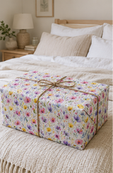 A square gift box wrapped in white wrapping paper with a colorful floral print and tied with a brown twine bow, placed on a beige knit blanket in a softly lit bedroom.