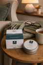 A tin of Muscle & Joint Balm sits on a wooden table next to its box, styled with a mug, glasses, and a throw blanket in a cozy living room setting.