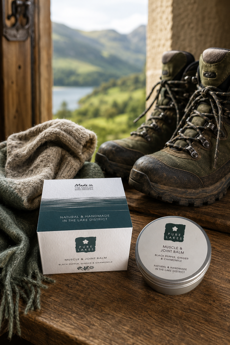 A tin of Muscle & Joint Balm and its outer box are displayed on a rustic wooden surface next to hiking boots and a knitted blanket, with a scenic mountain landscape in the background.