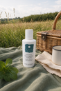 A bottle of Pure Lakes Natural Anti-Insect Lotion stands on a green blanket outdoors, styled with a wicker picnic basket, a white mug, mint leaves, and a grassy field in the background.
