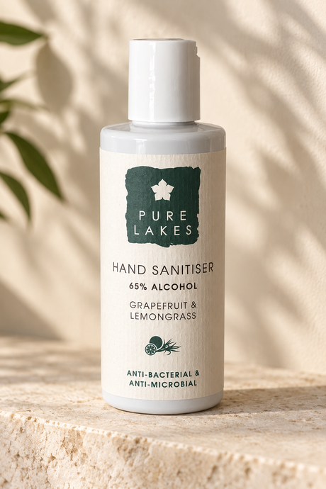 A bottle of Pure Lakes Hand Sanitiser, grapefruit and lemongrass scent, 65% alcohol, standing on a textured stone surface with soft leaf shadows cast over it.