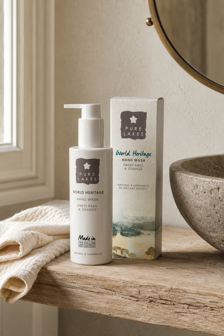Pure Lakes World Heritage Hand Wash bottle and box displayed on a rustic wooden shelf next to a folded cream towel, with a stone bowl and a round mirror in the background.