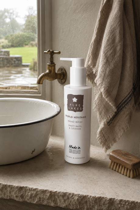 A bottle of Pure Lakes World Heritage Hand Wash stands on a stone sink ledge beside a white enamel basin, brass faucet, hanging towel, and a wooden scrub brush, with a window showing an outdoor garden view.