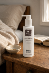 A bottle of Pure Lakes Sweet Basil & Orange Hand & Body Lotion stands on a rustic wooden bedside table in a softly lit bedroom, with a small bowl, open book, and bed in the background.