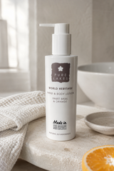 A bottle of Pure Lakes Hand & Body Lotion on a stone surface next to a folded textured towel and a slice of orange, in a bright, neutral bathroom setting.