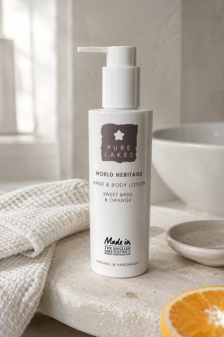 A bottle of Pure Lakes Hand & Body Lotion on a stone surface next to a folded textured towel and a slice of orange, in a bright, neutral bathroom setting.