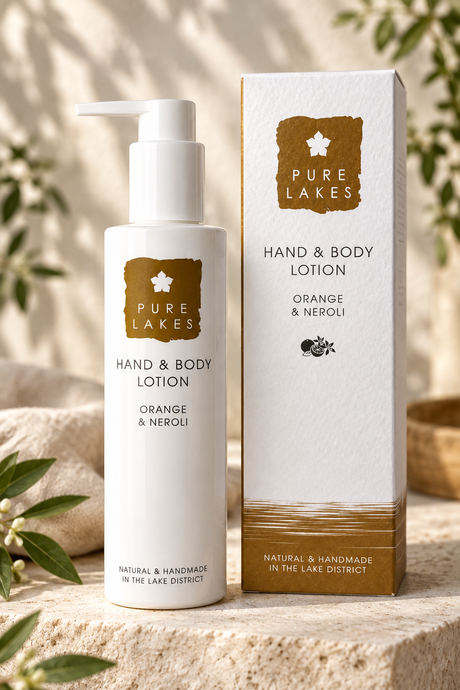 Pure Lakes Hand & Body Lotion in Orange & Neroli scent, shown as a white pump bottle next to its white and gold packaging box on a stone surface with soft botanical background.