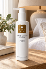 A bottle of Pure Lakes Hand & Body Lotion in Orange & Neroli scent, standing on a wooden bedside table in a softly lit bedroom setting.
