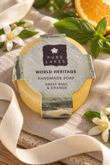A round bar of Pure Lakes Sweet Basil & Orange handmade soap wrapped in a decorative paper sleeve, styled on a beige linen cloth with basil leaves, white flowers, and an orange slice.