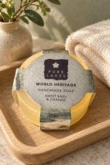 A round bar of Pure Lakes handmade soap labeled 'World Heritage Sweet Basil & Orange' resting on a wooden tray, with a beige towel and a small potted plant in the background.