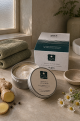 A tin of Muscle & Joint Balm with its lid placed in front, alongside a branded product box. The scene is styled on a stone surface with folded green towels, ginger root, chamomile flowers, and a small dish, suggesting a spa-like environment.