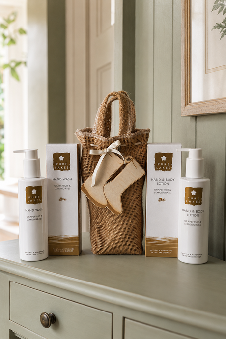 A hand and body care gift set from Pure Lakes arranged on a light sage green dresser. The set includes a gift bag with a cream ribbon and welly tag which can be personalised, two white pump bottles labeled Hand Wash and Hand & Body Lotion, and two matching white boxes, all in a warm, neutral-toned room.