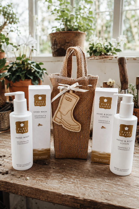 A set of Pure Lakes hand and body care products arranged on a rustic wooden table in front of a window with potted plants. The set includes a hand wash, a hand & body lotion bottle, a hand & body lotion box, a  gift bag with a boot-shaped tag, and a matching gift box.
