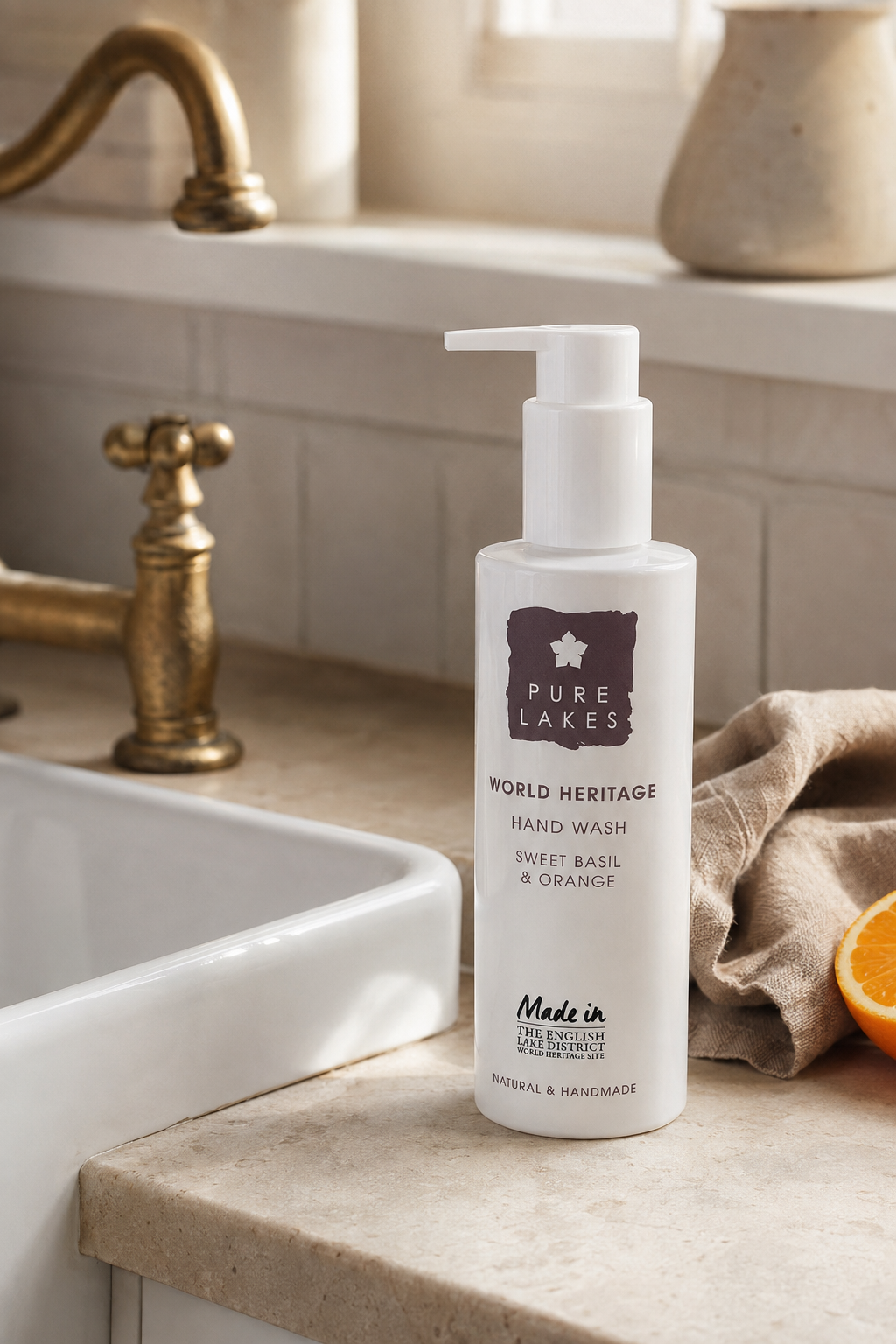 A bottle of Pure Lakes World Heritage Hand Wash, Sweet Basil & Orange, displayed on a beige stone countertop next to a white sink, a brass faucet, a beige cloth, and a slice of orange.