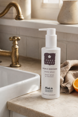 A bottle of Pure Lakes World Heritage Hand Wash, Sweet Basil & Orange, displayed on a beige stone countertop next to a white sink, a brass faucet, a beige cloth, and a slice of orange.