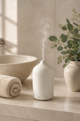 White diffuser emitting steam on a bathroom counter with a plant and towel in the background