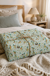 A rectangular gift box wrapped in a light blue-green Quirky Duck patterned paper  illustrations and tied with a twine bow, placed on a bed with beige bedding and a wooden headboard in a softly lit bedroom.