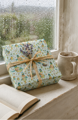 A wrapped gift box with a water colour quirky duck pattern and a raffia ribbon sits on a windowsill next to an open book and a ceramic mug. Raindrops are visible on the window, and a bouquet of dried lavender rests on top of the package.