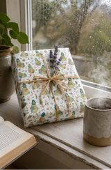 A gift box wrapped in white paper with a green and quirky duck pattern, tied with twine and topped with lavender sprigs, sitting on a windowsill next to an open book and a ceramic mug.