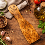 Wooden spatula with 'Top Chef' engraving on a wooden surface with tomatoes, oil, and herbs.