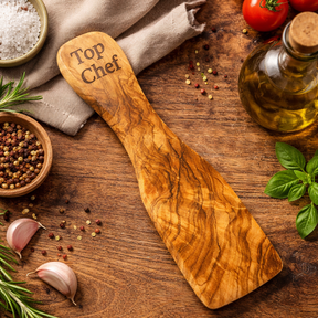 Wooden spatula with 'Top Chef' engraving on a wooden surface with tomatoes, oil, and herbs.