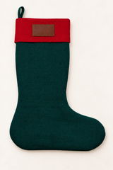 A linen Christmas stocking with a dark forest green body and a red cuff, featuring a small brown leather rectangular patch on the cuff and a hanging loop at the top, shown against a plain off-white background.