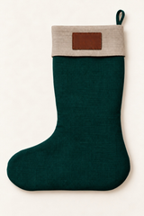 A dark green linen Christmas stocking with a light beige fabric cuff and a brown rectangular patch on the front, shown against a plain off-white background.