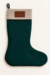 A linen Christmas stocking with a dark green body and a natural linen fabric cuff featuring a brown leather rectangular patch, shown against a plain off-white background.