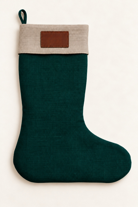 A linen Christmas stocking with a dark green body and a natural linen fabric cuff featuring a brown leather rectangular patch, shown against a plain off-white background.