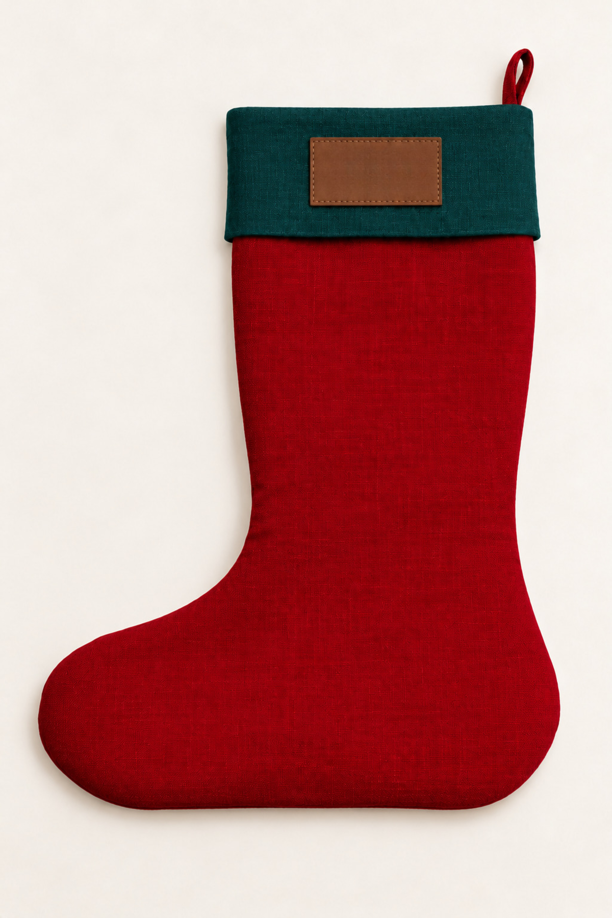 A red linen Christmas stocking with a dark green cuff and a brown leather rectangular patch, shown against a plain off-white background.