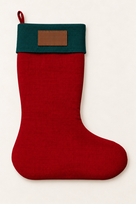 A festive linen Christmas stocking made of red linen, featuring a dark green cuff with a brown leather rectangular patch and a hanging loop at the top, shown against a plain off-white background.