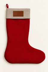 A red linen Christmas stocking with a natural colour fabric cuff and a brown rectangular leather patch on the front, shown against a plain off-white background.