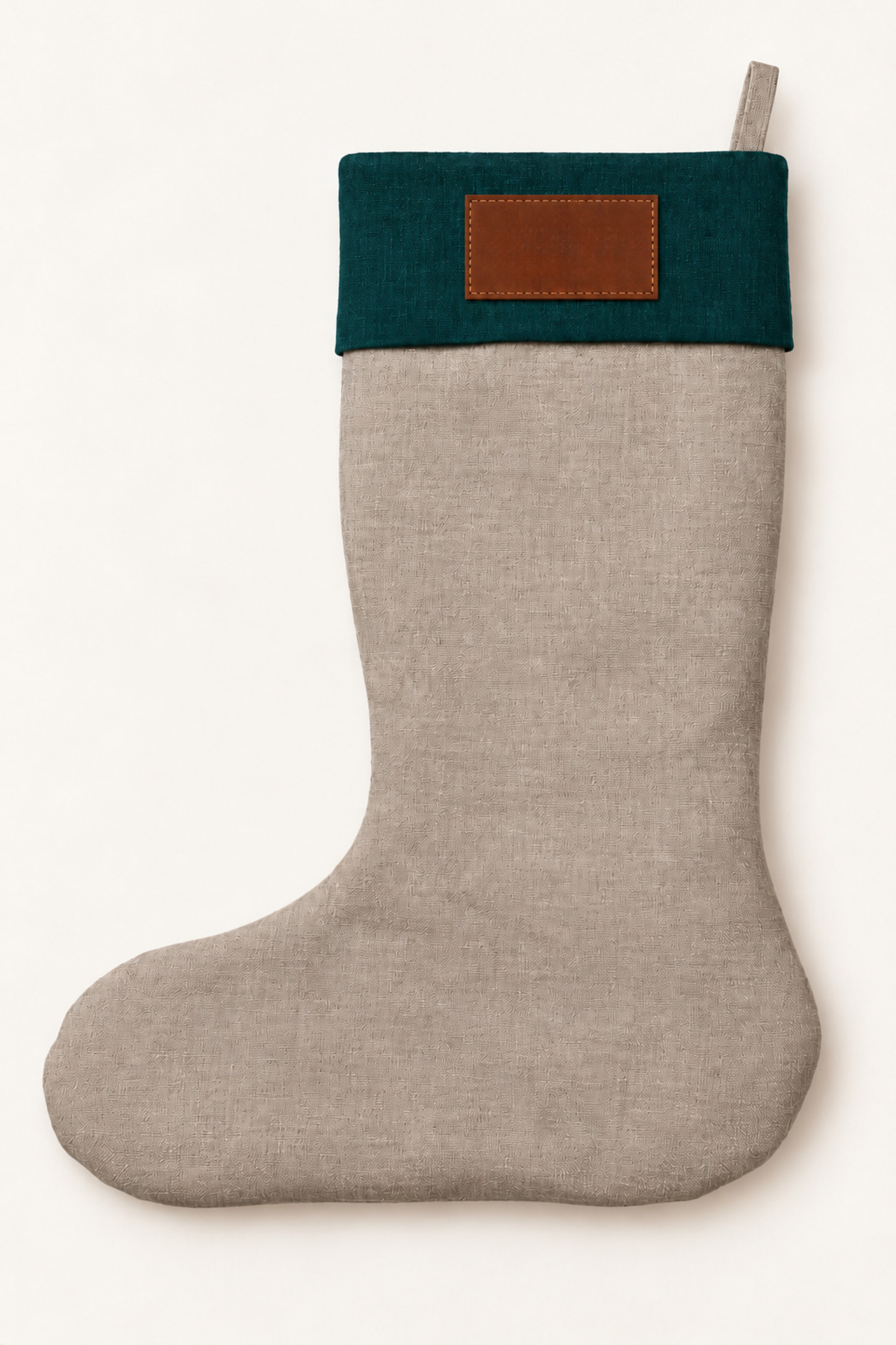 A light natural linen Christmas stocking with a dark forest green cuff and a brown leather patch on the front, shown against a plain off-white background.