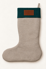 A light natural linen Christmas stocking with a dark forest green cuff and a brown leather patch on the front, shown against a plain off-white background.