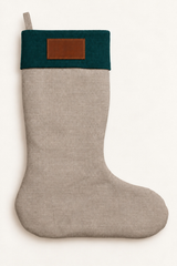 A light beige linen Christmas stocking with a dark teal green cuff and a brown leather patch on the front, shown against a plain off-white background.