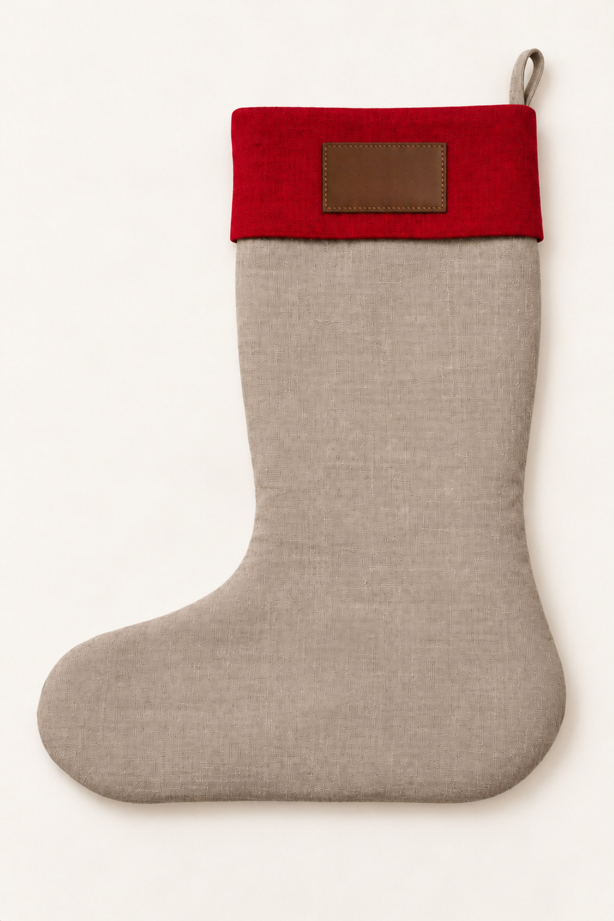 A linen Christmas stocking with a natural linen body and a vibrant red cuff, featuring a blank brown leather patch on the cuff and a hanging loop at the top, set against a plain off-white background.