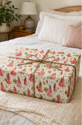 A rectangular gift box wrapped in cream wrapping paper with a repeating pattern of pink and red vintage dresses and roses, tied with a rustic brown twine bow, placed on a bed with white linens and a neutral bedroom background.