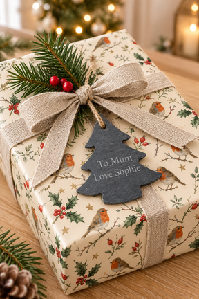 A wrapped Christmas gift box on a wooden surface, covered in cream wrapping paper with a robin and holly print, tied with a beige ribbon and bow, and decorated with a Christmas tree shaped slate gift tag that reads 'To Mum Love Sophie'.
