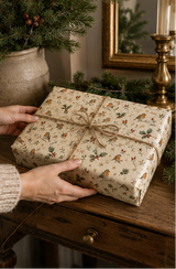 Christmas wrapping paper featuring red-breasted birds perched among holly leaves, red berries, rosehips, leafy sprigs, bare winter twigs and small golden stars. The illustrated repeat uses deep green, berry red, warm brown and soft grey on a warm cream background.