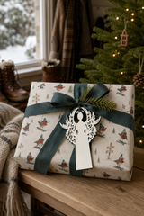 White filigree angel gift tag on a present wrapped in Robin Ski Derby Christmas paper beside a snowy window.