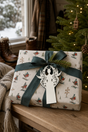White filigree angel gift tag on a present wrapped in Robin Ski Derby Christmas paper beside a snowy window.