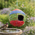 Colorful woven birdhouse hanging in a garden setting with flowers and plants.
