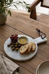 A round white marble serving board with a handle, styled on a rustic wooden table with sliced bread, grapes, olives, and a small bowl of dip. A beige napkin and a vase with greenery are also visible in the softly lit setting.