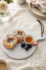A round white marble serving board with a handle, styled with two golden-brown pastry cookies, a small bowl of honey, blackberries, and a strawberry on a beige linen tablecloth.