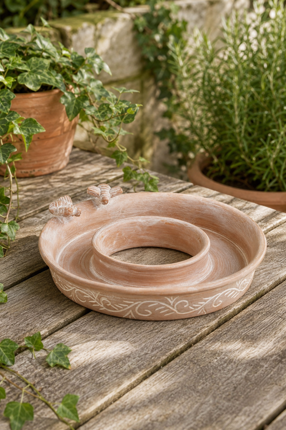 A terracotta garden bird feeder, bird bath with a central ring and decorative scrollwork, displayed on a weathered wooden table with potted plants in the background.