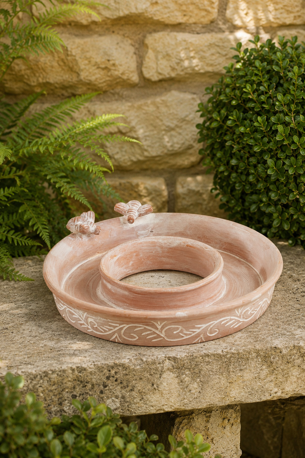 A terracotta outdoor garden bird feeder and bird bath with two small decorative bees and a floral pattern around the rim, set on a stone ledge with green plants and a stone wall in the background.