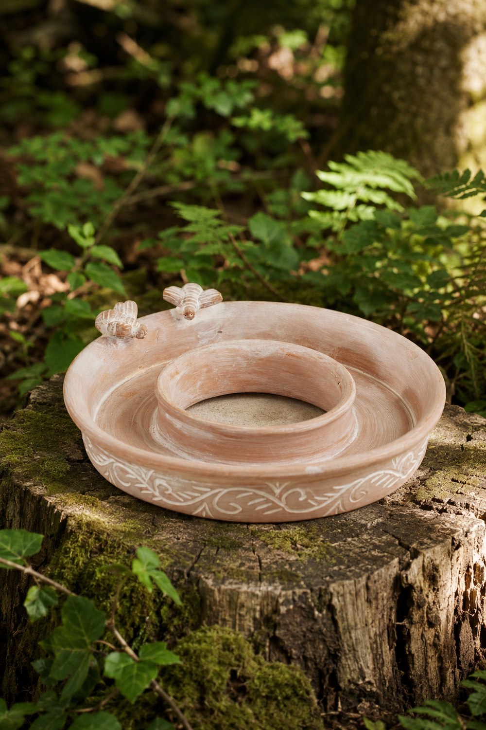 A terracotta bird feeder and bird bath with a central well and decorative floral details, displayed on a mossy tree stump in a lush green outdoor setting.