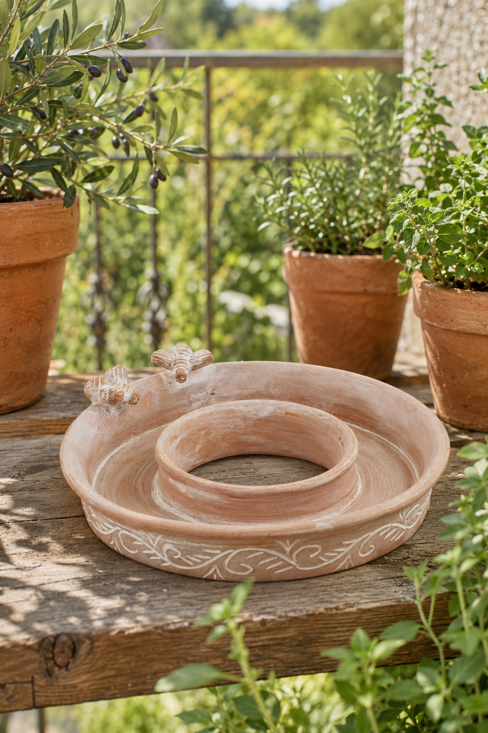 A terracotta garden bird feeder and bird bath with a central ring two bees and decorative scrollwork, displayed on a rustic wooden table outdoors with potted plants in the background.